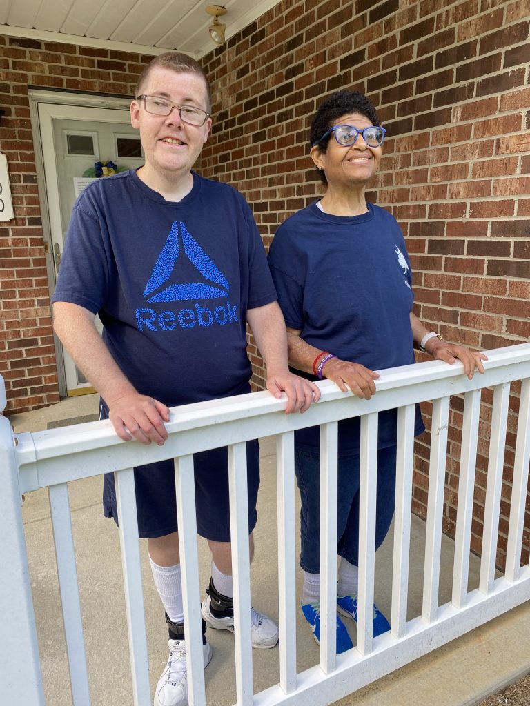 Two individuals stand together on a porch, smiling and leaning on a white railing. They are dressed in navy blue shirts and shorts, with one wearing glasses and the other wearing braces on their legs. A brick house serves as the backdrop.