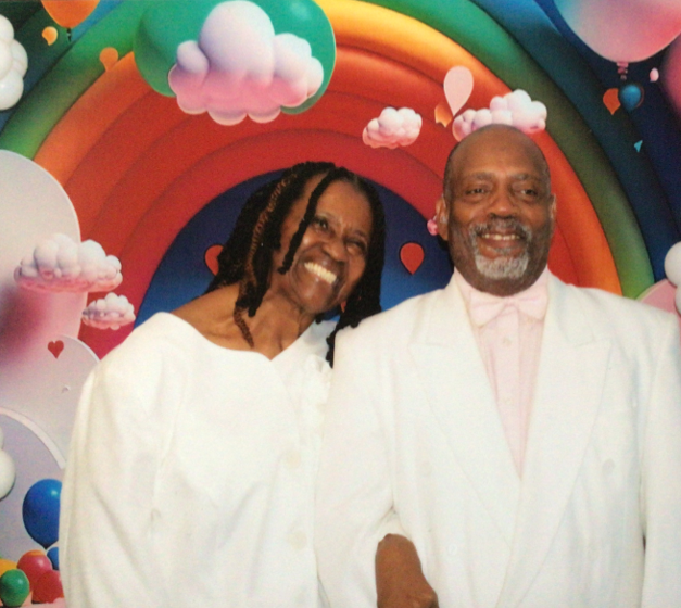 Couple smiling in white outfits with a rainbow background.