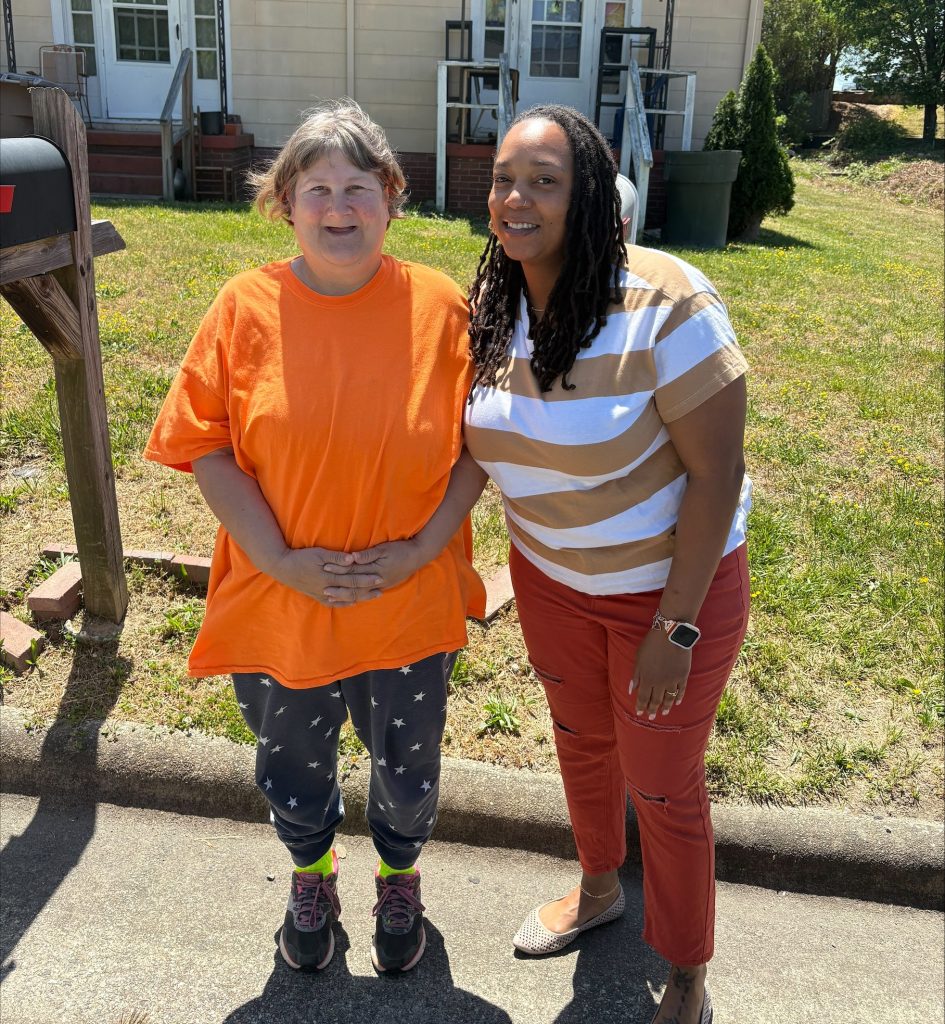 Two women standing together outside and smiling. On the left is Kristine Maher, wearing an orange shirt. On the right is Angelique Graham, wearing a brown and white shirt.