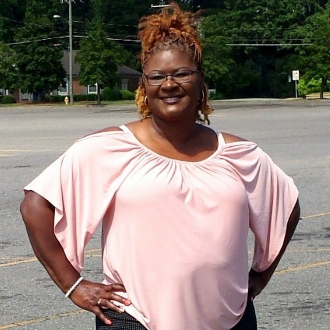 Essie McMillan stands with her hands on her hips and a huge smile on her face dressed in a light color blouse, black pants and sandals.