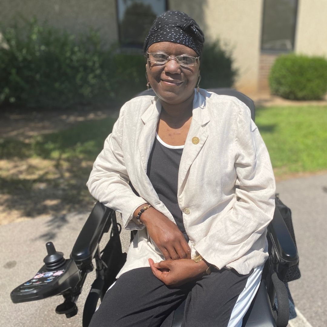 Kashinda Marche, in white jacket and black top and pants, sits in her wheelchair in front of the Monarch Behavioral Health Outpatient Clinic in Raleigh.