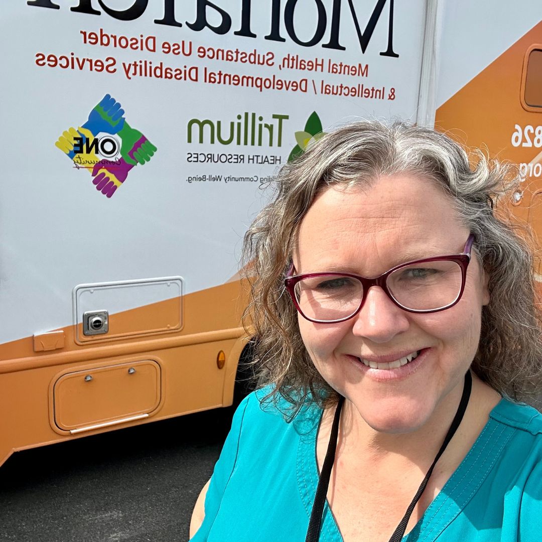 Registered Nurse Kelly Hagwoodl in front of Monarch's Mobile Integrated Care Clinic dressed a turquoise scrub shirt, wearing glasses and smiling. 